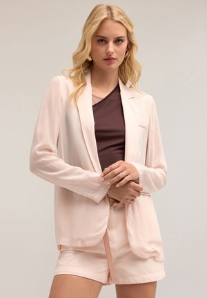 Lichtroze blazer van lichtgewicht stof, met een enkele knoopsluiting, lange mouwen en een losse pasvorm, gedragen over een bruine top.