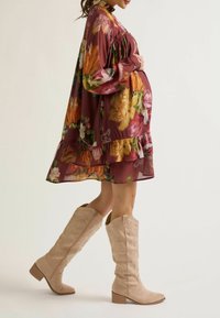 Zwangere vrouw in een bloemenjurk die haar buik omarmt, met hoge beige laarzen aan. Ze staat in profiel tegen een lichte achtergrond.