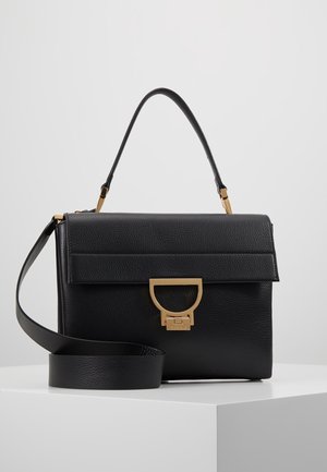 Sort tekstureret lædertaske med håndtag øverst, aftagelig skulderrem og gyldent D-formet lukke foran på hvidt udstillingsbord.