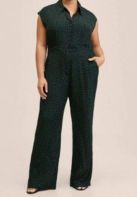 Žena ve černém bezramenném overal s zelenými puntíky, na knoflíky vpředu a širokými nohavicemi, s jednou rukou v kapse.