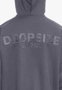 Hanorac gri cu text ridicat „DROPSIZE” și „EST.2020” imprimat pe spate. Materialul are o textură netedă și o croială casuală.
