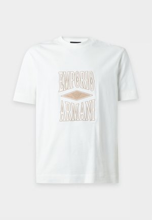 Ung mand står iført hvid Emporio Armani T-shirt, sorte bukser med hvide striber og hvide sneakers mod en ensartet lys baggrund.