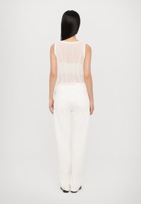 Balts bez piedurknēm adīts tops ar vertikāliem svītru rakstiem, kam ir noapaļots aizmugure; apvienots ar vaļīgām baltām biksēm un melnām kurpēm.