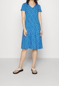 Femme portant une robe à fleurs bleues longueur genou et des sandales à plateforme beige, se tenant devant un fond blanc uni.
