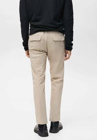 Beige trousers made of smooth fabric, featuring two back pockets and a tapered fit, paired with black shoes from a low-angle perspective.