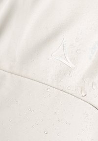 Light beige fabric with a smooth texture, featuring water droplets and a subtle logo in the corner, indicating water-resistant properties.