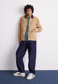 Chaqueta de lona color arena con un cuello marrón, botones dorados y dos bolsillos frontales; combinada con una camiseta color azul verdoso y pantalones de pierna ancha azul oscuro.