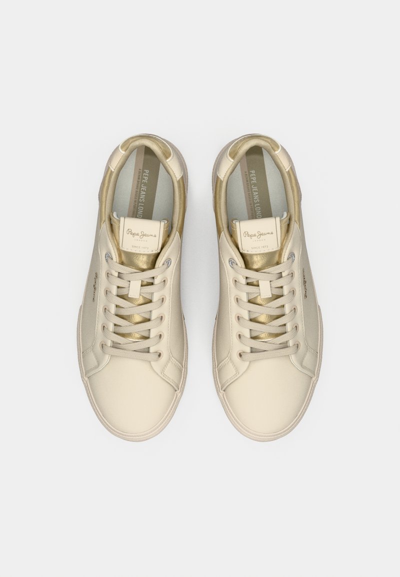 Zapatillas beige con parte superior de cuero suave, detalles dorados, cordones planos beige y plantilla acolchada. Etiqueta de la marca visible en la lengüeta.