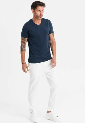 Marineblaues V-Ausschnitt-T-Shirt aus weichem Stoff, kombiniert mit weißen, schmalen Hosen und weißen Sneakern. Auffällige Tattoos an den Armen; neutraler Hintergrund.