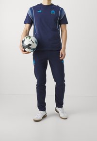 Person in blue Puma sportswear holding a white-black Puma soccer ball, standing against a plain light background.