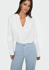 Femme portant un chemisier blanc à manches longues avec des détails en dentelle et un pantalon bleu clair taille haute, posant devant un fond uni.