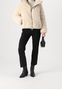 Cremefarbige Kunstfelljacke mit Reißverschluss vorne und Kapuze, kombiniert mit schwarzen, maßgeschneiderten Hosen und schwarzen, hochhackigen Stiefeletten. Kleine schwarze Handtasche.