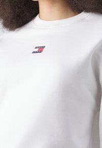 Primo piano di una persona che indossa una felpa bianca con colletto girocollo, con un piccolo logo Tommy Jeans sul petto.