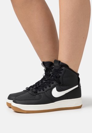 Baskets montantes noires Nike avec swoosh blanc et semelles en gomme, portées sur des jambes nues, sur un fond blanc.