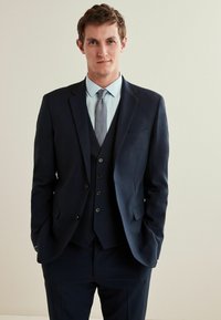 Traje de tres piezas azul marino con solapa pico, pañuelo de bolsillo y botones. Una camisa azul clara y una corbata estampada completan el look formal.