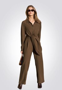 Bruine jumpsuit met lange mouwen en een strik in de taille, met een knoopsluiting aan de voorkant, wijde pijpen, en gecombineerd met een paar donkere zonnebrillen en een handtas.