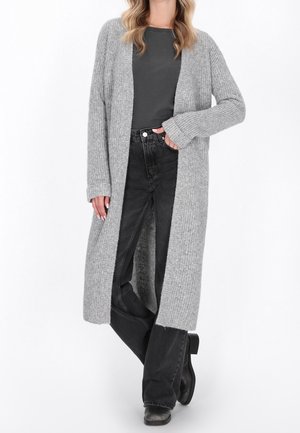Mujer que lleva un largo cárdigan de punto gris, camiseta gris oscuro, vaqueros negros y zapatos negros, de pie con una mano en el bolsillo.
