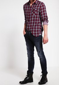 Camicia a maniche lunghe a quadri rossi e blu con tasca sul petto, abbinata a jeans in denim scuro e stivali neri con lacci, che mostra un look casual.