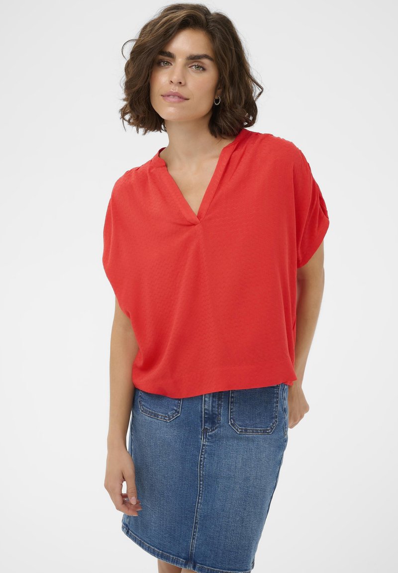 Femme aux cheveux bruns courts portant un chemisier rouge à col en V et une jupe en jean bleue mi-longue, debout devant un fond blanc.