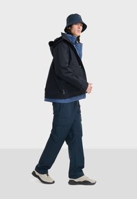 Veste imperméable marine avec capuche, superposée à une chemise bleue ; associée à un pantalon cargo bleu et des chaussures de randonnée beiges ; chapeau seau noir.