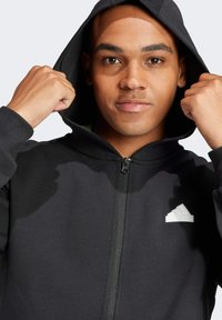 Felpa nera con zip realizzata in materiale morbido, con cappuccio, zip frontale e logo Adidas bianco sul lato sinistro del petto.