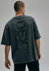 Szary, oversize t-shirt z teksturowanym czarnym graficznym nadrukiem "DEXT" w pionie na plecach. Krótkie rękawy i luźny krój.