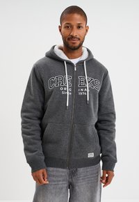 Hombre con una sudadera con cremallera de color gris oscuro, forrada de polar blanco, con el texto "CREEKS ORIGINAL Since 1974" y jeans de color gris claro, apoyado contra un fondo blanco.