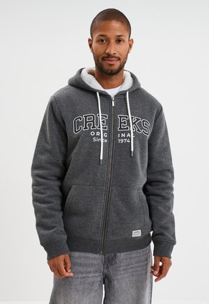 Hombre con una sudadera con cremallera de color gris oscuro, forrada de polar blanco, con el texto "CREEKS ORIGINAL Since 1974" y jeans de color gris claro, apoyado contra un fondo blanco.