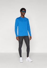 Zils garo piedurkņu sporta krekls ar ceturtdaļas rāvējslēdzēju, apvienots ar melnajām leggēm un baltajiem skriešanas apaviem. Uz krūtīm ir logotips.