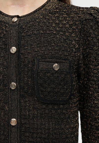 Μαύρο ένδυμα με υφή και λαμπερές χρυσές λεπτομέρειες, ασημένια διακοσμητικά κουμπιά κατά μήκος του μπροστινού μέρους και σε μια μικρή τσέπη στο στήθος.