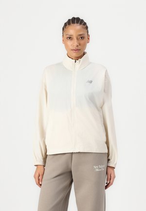 Lengvas, kreminės spalvos zip-up švarkas su stovinčiu apykakle, elastingomis rankovėmis ir atspindinčiu logotipu ant krūtinės; lygus paviršius, laisvas fasonas.