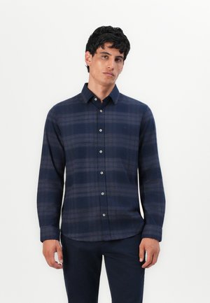 Man in een donkerblauw geruite overhemd met knopen en donkere broek, staande tegen een effen witte achtergrond.