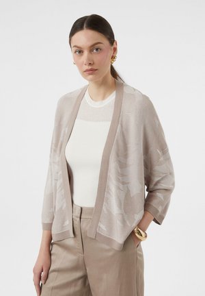 Lichtbeige cardigan met een gestructureerd breipatroon en een open voorkant, gedragen over een witte top, gecombineerd met beige broeken en accessoires.