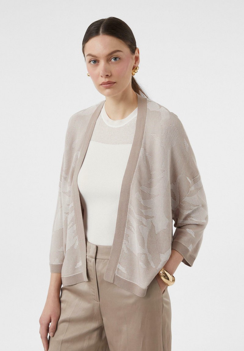 Jasno beżowy kardigan z teksturowanym splotem, z otwartym przodem, noszony na białej bluzce, w połączeniu z beżowymi spodniami i akcesoriami.