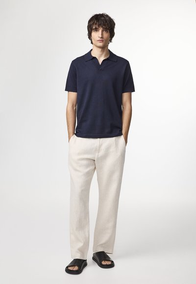 Männliches Modell steht mit den Händen in den Taschen und trägt ein marineblaues Polo mit kurzen Ärmeln, hellbeige Hose und schwarze offene Sandalen vor weißem Hintergrund.