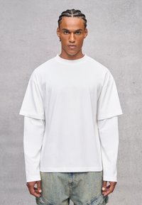 Weißes, übergroßes T-Shirt mit kurzen Ärmeln, das über einem Langarmshirt getragen wird. Baumwollmaterial, einfacher Rundhalsausschnitt, lockere Passform.
