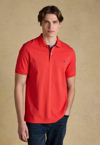 Jeune homme debout, portant un polo à manches courtes rouge vif et un jean bleu foncé, sur un fond beige uni.