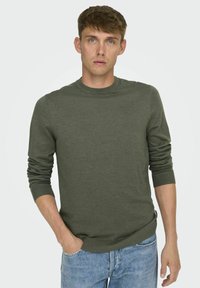Pull olive green, col rond, manches longues avec finitions côtelées, texture douce, associé à un jean bleu clair, fond uni.