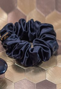 Czarny satynowy scrunchie o zbiegającej się fakturze. Widoczna mała metka z logo. Umieszczony na złotej powierzchni o heksagonalnym wzorze.