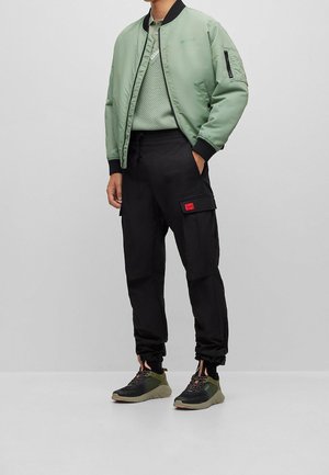 Homme portant une veste aviateur vert clair, un pull vert texturé assorti, un pantalon cargo noir avec un patch rouge, et des baskets noir-vert.