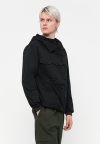 Black lightweight hooded jacket with a front pocket, drawstring at the waist, and buttoned chest pockets. Paired with olive green pants.