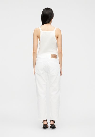 Άτομο με μακριά σκουρόχρωμα μαλλιά που φοράει λευκό αμάνικο τοπ, λευκά τζιν με δερμάτινο ταμπελάκι και μαύρα σανδάλια με ψηλό τακούνι, γυρισμένο προς τα πίσω.