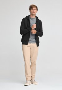 Sudadera negra con cremallera sobre una camiseta gris, combinada con pantalones beige claros y zapatillas beige. La sudadera tiene un diseño minimalista y cordones.