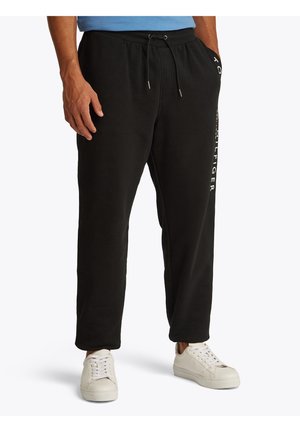 Mężczyzna ubrany w czarne spodnie dresowe Tommy Hilfiger z bocznym logo i białe sneakersy, stojący z jedną ręką w kieszeni.