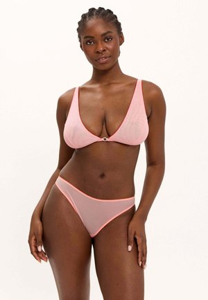Frau trägt einen hellrosa Netz-Bikini mit dünnen Trägern, steht vor einem schlichten weißen Hintergrund, blickt mit neutralem Ausdruck nach vorne.