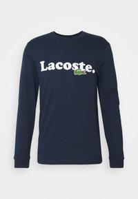 T-shirt à manches longues bleu marine en coton avec le logo "Lacoste" en blanc, et un accent de crocodile vert brodé sur le côté gauche.
