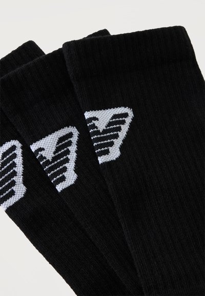 Trois chaussettes noires côtelées avec un logo triangulaire blanc comportant des rayures noires diagonales près du haut.