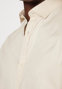 Light beige button-up shirt with a soft texture, featuring a pointed collar and subtle stitching details. Buttons are visible at the neck.