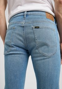 Mann trägt hellblaue Jeans mit Gesäßtaschen und Leder Markenetikett, kombiniert mit einem weißen, in die Hose gesteckten Hemd.
