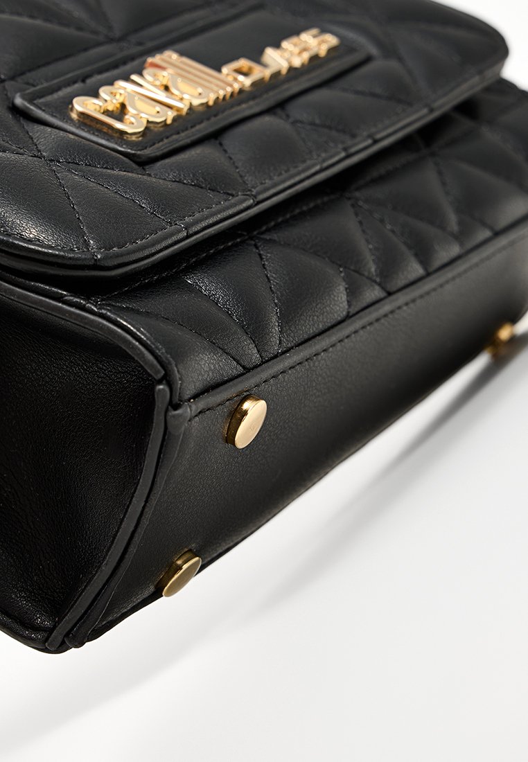 Bolso de cuero negro acolchado con acentos en hardware dorado y cierre de cremallera. Presenta una placa con el logo y pies de metal en la base para mayor estabilidad.